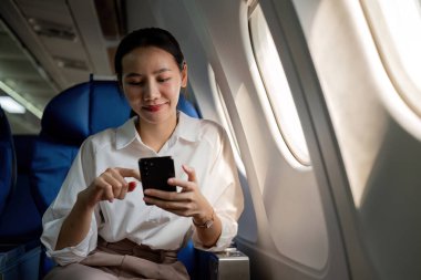 Genç Asyalı kadın haber bilgilerini kontrol etmek için cep telefonu kullanıyor. Business class 'ta pencerenin yanında, uçuş sırasında uçak dersi, seyahat ve iş konsepti..