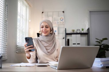 İşte tesettürlü mutlu Müslüman iş kadını ofiste finans ve pazarlama üzerine çalışmak için bilgisayarınızı ve akıllı telefonunuzu kullanın...