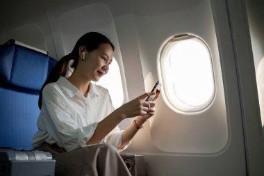 Genç Asyalı kadın haber bilgilerini kontrol etmek için cep telefonu kullanıyor. Business class 'ta pencerenin yanında, uçuş sırasında uçak dersi, seyahat ve iş konsepti..