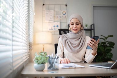 İşte tesettürlü mutlu Müslüman iş kadını ofiste finans ve pazarlama üzerine çalışmak için bilgisayarınızı ve akıllı telefonunuzu kullanın...
