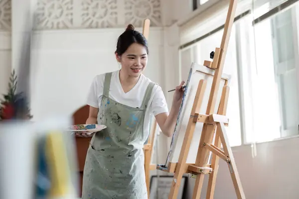 stock image Young Asian female artist is intently painting acrylic paints on canvas in a painting studio..