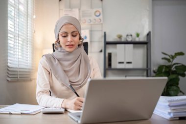 Günümüz iş yerinde çalışan Müslüman iş kadını bilgisayarını kullanarak belgelere not alıyor..