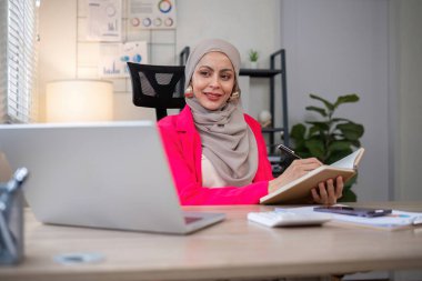 Günümüz iş yerinde çalışan Müslüman iş kadını bilgisayarını kullanarak belgelere not alıyor..