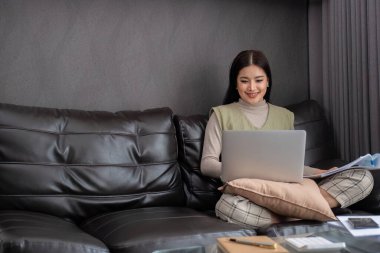 Orta yaşlı bir kadın yemek masasında dizüstü bilgisayarı ve yanında bir yığın belgeyle çalışıyor...