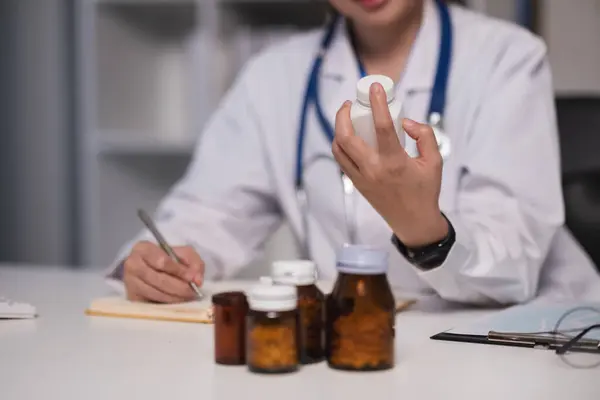 A woman doctor is holding a bottle of medicine in her hand. She is writing in a notebook