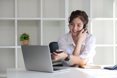 İş kadını beyaz kadın telefonda konuşuyor ve modern ofiste otururken gülümseyerek dizüstü bilgisayar kullanıyor..