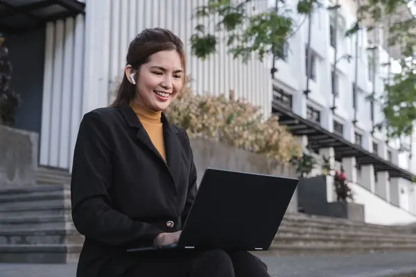 一个穿着黑色西服的女人坐在台阶上 面前有一台笔记本电脑 她面带微笑 乐在其中 — 图库照片