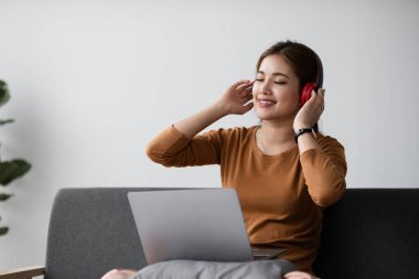 Genç Asyalı kadın boş zamanlarını kanepede oturup dinlenmek ve eğlenmek için kulaklık takarak müzik dinleyerek geçiriyor...