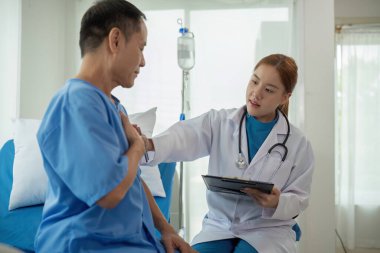 Bir doktor hastane odasında bir hastayla konuşuyor. Hasta mavi bir hastane önlüğü giyiyor.