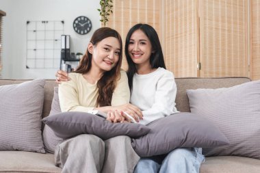 Genç LGBT kadını, lezbiyen çift, kanepede oturuyor, kucaklaşıyorlar ve birlikte mutlu bir şekilde gülümsüyorlar...