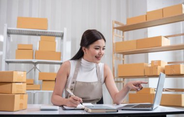 Bir kadın, çevrimiçi satmak için kutu kutu mal paketliyor, raflı ve dizüstü bilgisayarlı modern bir çalışma alanında çalışıyor, e-ticaret ve küçük işletmeler sergiliyor..