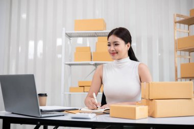 Bir kadın çevrimiçi satmak için kutu kutu mal paketliyor, paketler ve dizüstü bilgisayarla modern bir ev ofisinde çalışıyor..