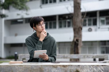 Açık havada okuyan genç bir üniversite öğrencisi, defter ve kalemle fikirler düşünüyor, modern kampüs geçmişi, eğitim ve öğrenim kavramı.