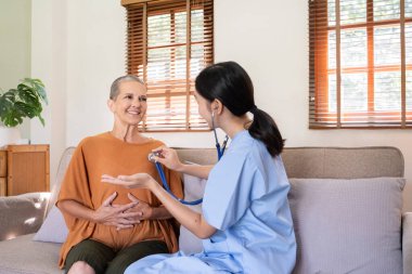 Mavi önlüklü bir bakıcı evde gülümseyen yaşlı bir kadının sağlığını kontrol etmek için steteskop kullanır..