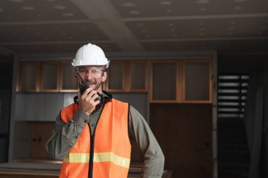 Young engineer at a construction site, wearing safety gear and using a radio for communication, in a modern building under construction. clipart