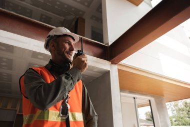 Genç bir mühendis bir inşaat alanında telsiz kullanıyor, güvenlik ekipmanı ve yansıtıcı yelek giyiyor, modern bir bina projesini denetliyor..