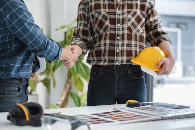 Modern bir ofiste iki erkek mühendis el sıkışıyor, planlar ve güvenlik teçhizatlarıyla bir inşaat projesinde işbirliği yapıyor..