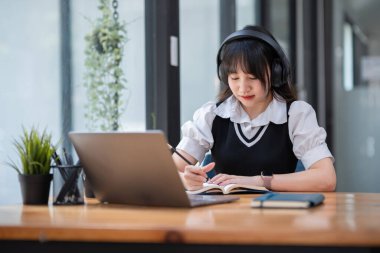 Laptop ve defterle çalışan, kulaklık takan genç bir üniversite öğrencisi, modern kampüs ortamında..