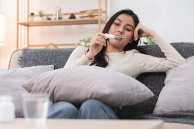 Termometre tutan genç bir kadın, endişeli görünüyor, elinde yastıkla kanepede oturuyor, sağlık veya hastalık belirtileri gösteriyor..