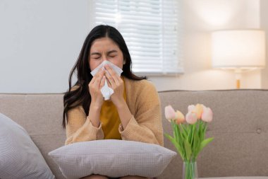 Oturma odasında rahat bir kanepede laleler ve arka planda bir lambayla oturan bir kadın mendile hapşırıyor..