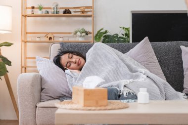 Kendini rahatsız hisseden bir kadın kanepeye kıvrılır, battaniyeye sarınır, rahat bir oturma odasında, masanın üzerinde bir mendil kutusu ve ilaçla..