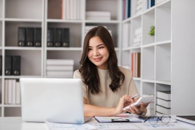 Evde çalışan genç bir muhasebeci, modern bir ofis ortamında dizüstü bilgisayar ve hesap makinesi kullanıyor..