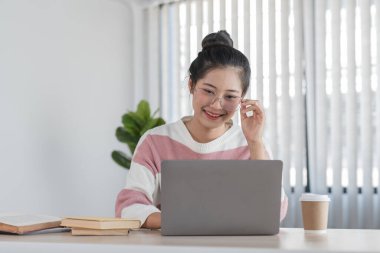 Evde internetten ders çalışan genç bir kadın. Bilgisayarıyla gülümsüyor ve meşgul. Etrafı kitaplar ve kahve fincanlarıyla çevrili..