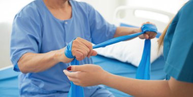 Hastanede yatan yaşlı bir hasta direnç bantları kullanan genç bir kadın terapistten fizik tedavi görüyor..