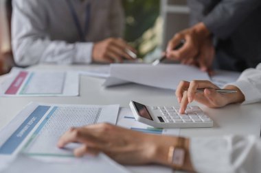 İş kadınlarının elleri, profesyonel ofis ortamında iş belgeleriyle çevrili bir hesap makinesiyle finansal hesaplamalar yapıyor..