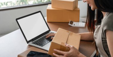 Bir kadın çevrimiçi satışlar için ürünleri paketlerken modern çalışma alanında beyaz ekranlı bir laptop kullanıyor, kutular ve akıllı telefonlarla çevrili..