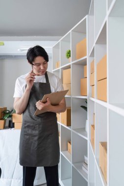 Önlüklü genç bir girişimci modern bir ev ofisinde çevrimiçi satış envanterini yönetiyor. Etrafı ürün ve nakliye kutularıyla çevrili..