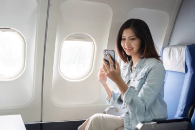 Genç bir iş kadını uçak penceresinin yanında oturuyor, akıllı telefonunu kullanıyor ve gülümsüyor, gündüz uçuşunda sıradan bir kot ceket giyiyor..