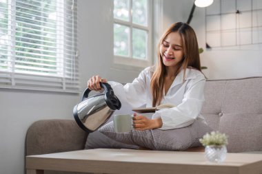 Bir kadın sabah rutini sırasında parlak, rahat bir oturma odasında kitap okurken çaydanlıktan bardağa kahve doldurur..