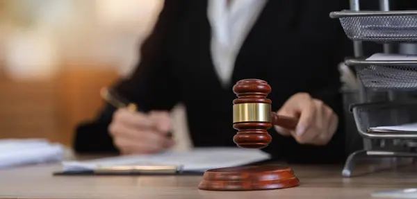 Bir kadın avukat, bir ofis ortamında açılan davayla ilgili sözleşme belgelerini incelerken masanın üzerine belirgin bir tokmak yerleştirilir..
