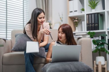 İki genç kadın evde birlikte ders çalışıyor, bir dizüstü bilgisayar ve defterle sınavlara hazırlanmak için birbirlerine yardım ediyorlar..
