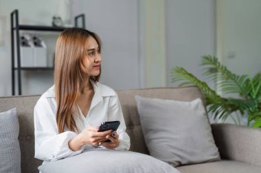 Bir kadın elinde cep telefonuyla kanepede oturuyor. Yüzünde ciddi bir ifadeyle telefona bakıyor.