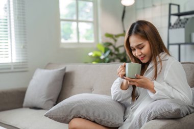 Bir kadın sabah sıcak bir fincan çayla doğal ışık ve rahat mobilyalarla dolu sıcak bir oturma odasında rahatlar..