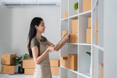 Önlüklü genç bir kadın modern bir depoda ürün stoklarını kontrol ediyor. Raflardaki envanteri yönetmek için bir pano kullanıyor..