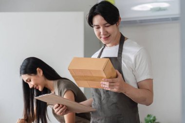 Neşeli bir çift, evlerinde çalışan online işlerinde ürün paketliyor ve bir pano ve bir kutuyla siparişleri yönetiyor..