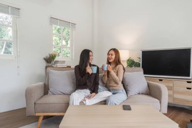 Lezbiyen çift rahat bir kanepede oturuyor, kahveyi paylaşıyor ve modern bir oturma odasında gülümsüyor..