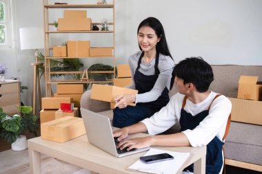 Çevrimiçi ürün satmak için işbirliği yapan bir çift, paketleme kutularıyla çevrili, modern bir ev ortamında dizüstü bilgisayar kullanıyor..