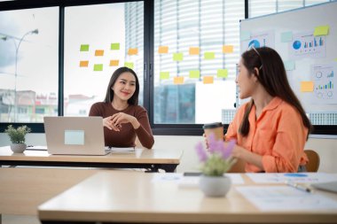 İki genç kadın muhasebeci modern bir ofiste iş hakkında konuşuyor. Dizüstü bilgisayarları, çizelgeleri ve penceresinde yapışkan notları var..