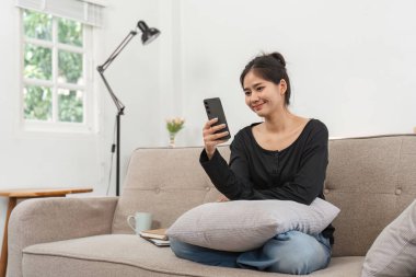 Kanepede rahat bir şekilde oturan bir kadın, akıllı telefonunu son gelişmelerden haberdar olmak için kullanıyor. Modern, aydınlık bir oturma odasında..