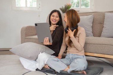 A joyful lesbian couple sitting on the floor in a cozy living room, sharing a moment of laughter while using a tablet and headphones. clipart