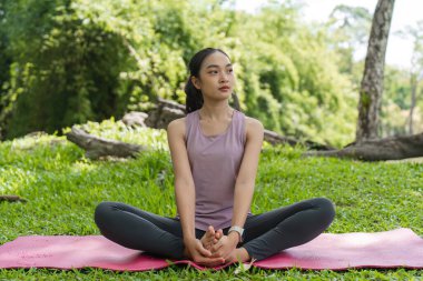 Huzur parkında yoga yapan bir kadın, pembe bir paspasın üzerinde oturuyor, yemyeşil, huzurlu bir anın tadını çıkarıyor..