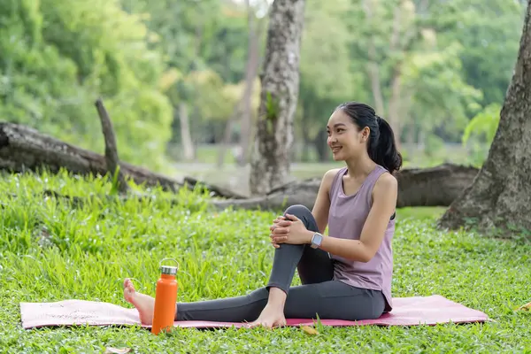 Bir kadın, doğa ve ağaçlarla çevrili yemyeşil bir parktaki paspasın üzerinde huzur dolu bir yoga seansından zevk alır. Rahatlama ve refahı teşvik eder..