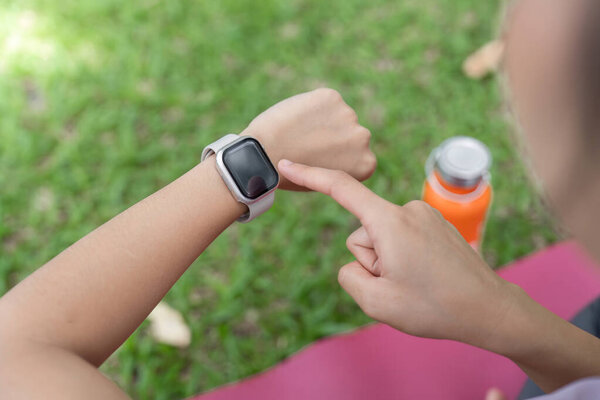 A young healthy woman checking her smartwatch while exercising outdoors, promoting fitness and an active lifestyle in a natural setting.