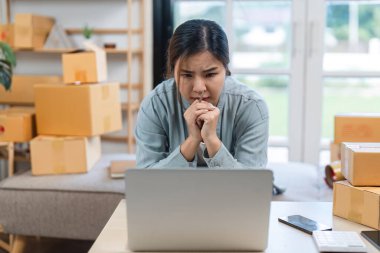 Genç bir kadın, çevrimiçi satış işini evden yönetirken stresli görünüyor. Etrafı kargo kutularıyla çevrili ve dizüstü bilgisayarı üzerinde çalışıyor..