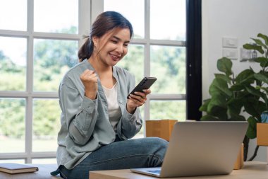 Genç bir kadın online alışveriş işinin başarısını modern bir ofis ortamında dizüstü bilgisayar ve akıllı telefon kullanarak sevinçle kutluyor..