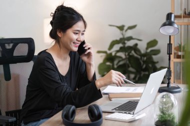 Evden çalışan genç bir kadın muhasebeci, modern bir ofis ortamında dizüstü bilgisayarı ve telefonu ile birden fazla iş yapıyor..